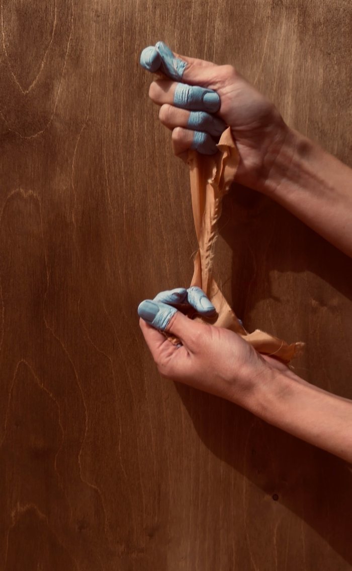 Zwei Hände mit blau gefärbten Fingerkuppen ziehen ein terrakottafarbenes Tuch auseinander.