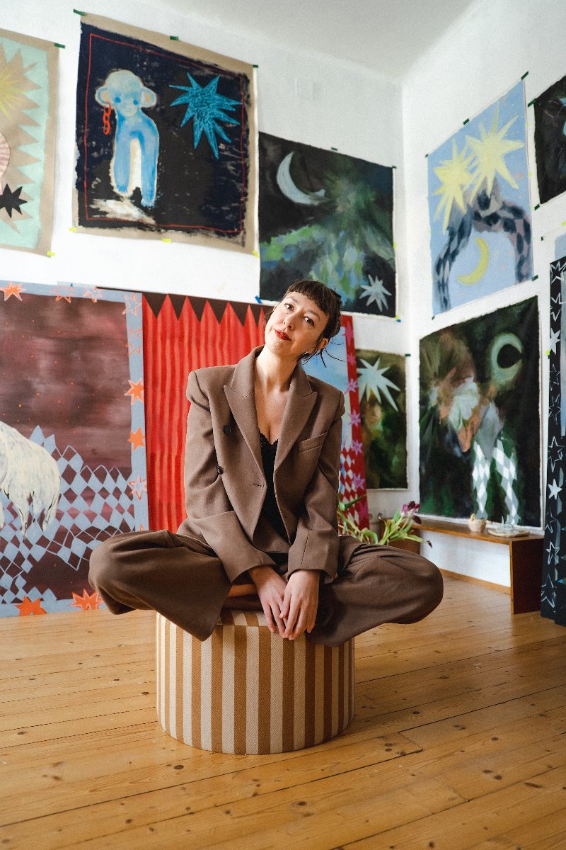 Theresa Gregor sitzt im Schneidersitz auf einem gestreiften Hocker in ihrem Atelier, umgeben von großformatigen Bildern mit Sternmotiven.